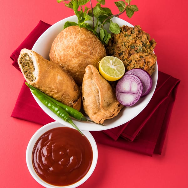 A white plate of Pakistani snacks including samosas, pakoras, and kachori, with chili, onion, and lime.