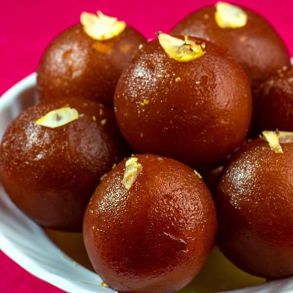 A white bowl filled with several glistening, dark brown gulab jamun, garnished with slivered almonds.