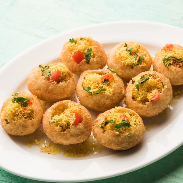 A white plate holds several pani puri, filled with yellow sev, diced tomatoes, and cilantro.