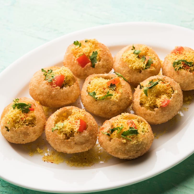 A white plate holds several pani puri, filled with yellow sev, diced tomatoes, and cilantro.
